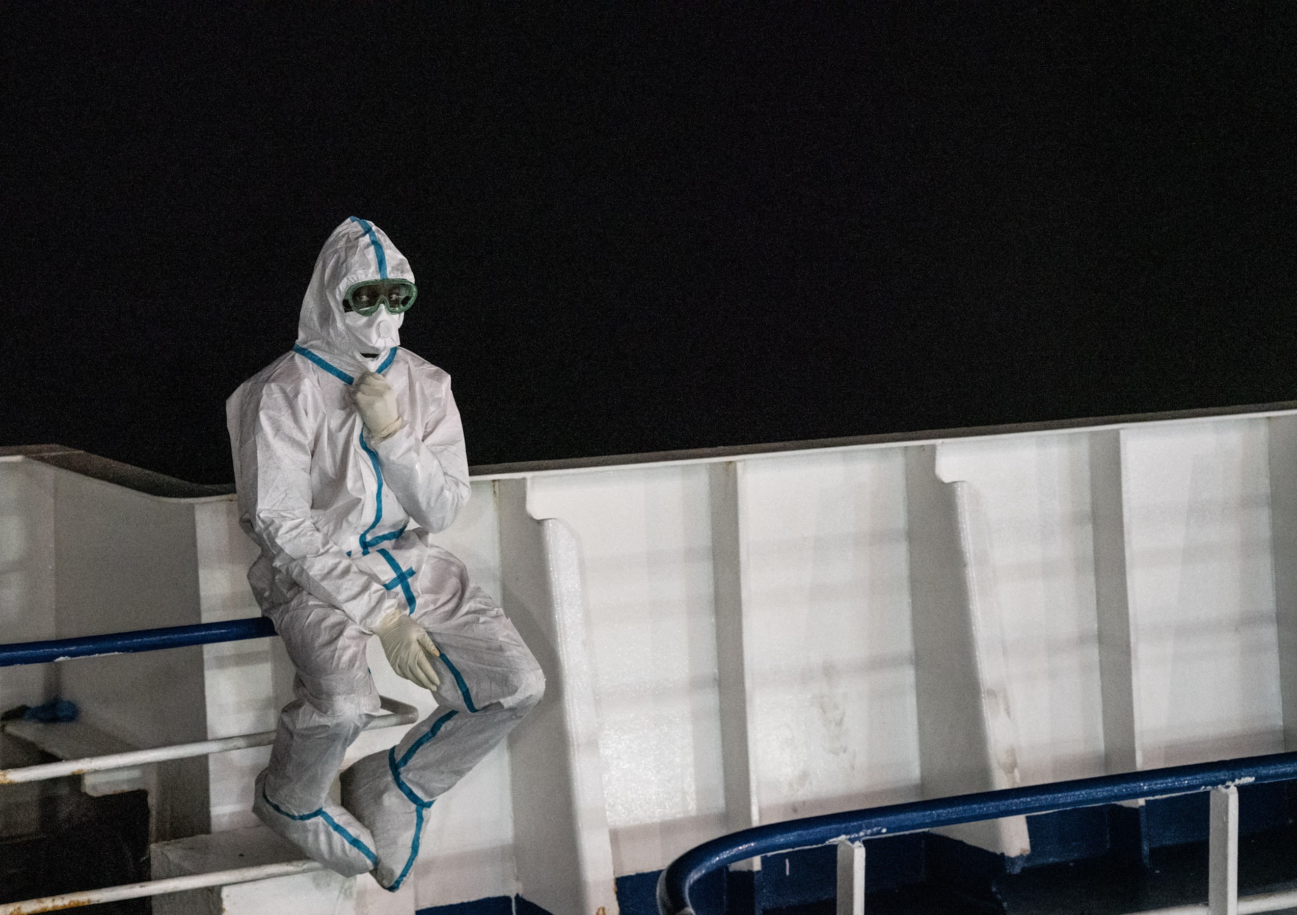 A Red Cross worker in PPE waits on one of the ships at night